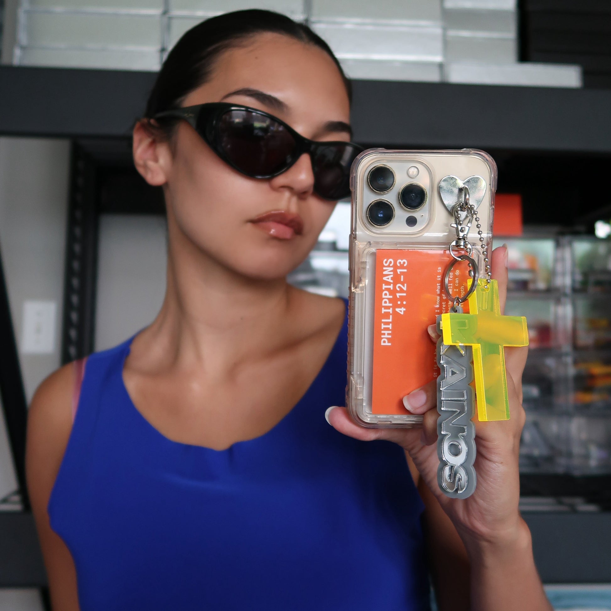Woman holding a phone case with a Kainos keychain, cross keychain, and scripture, wearing sunglasses.