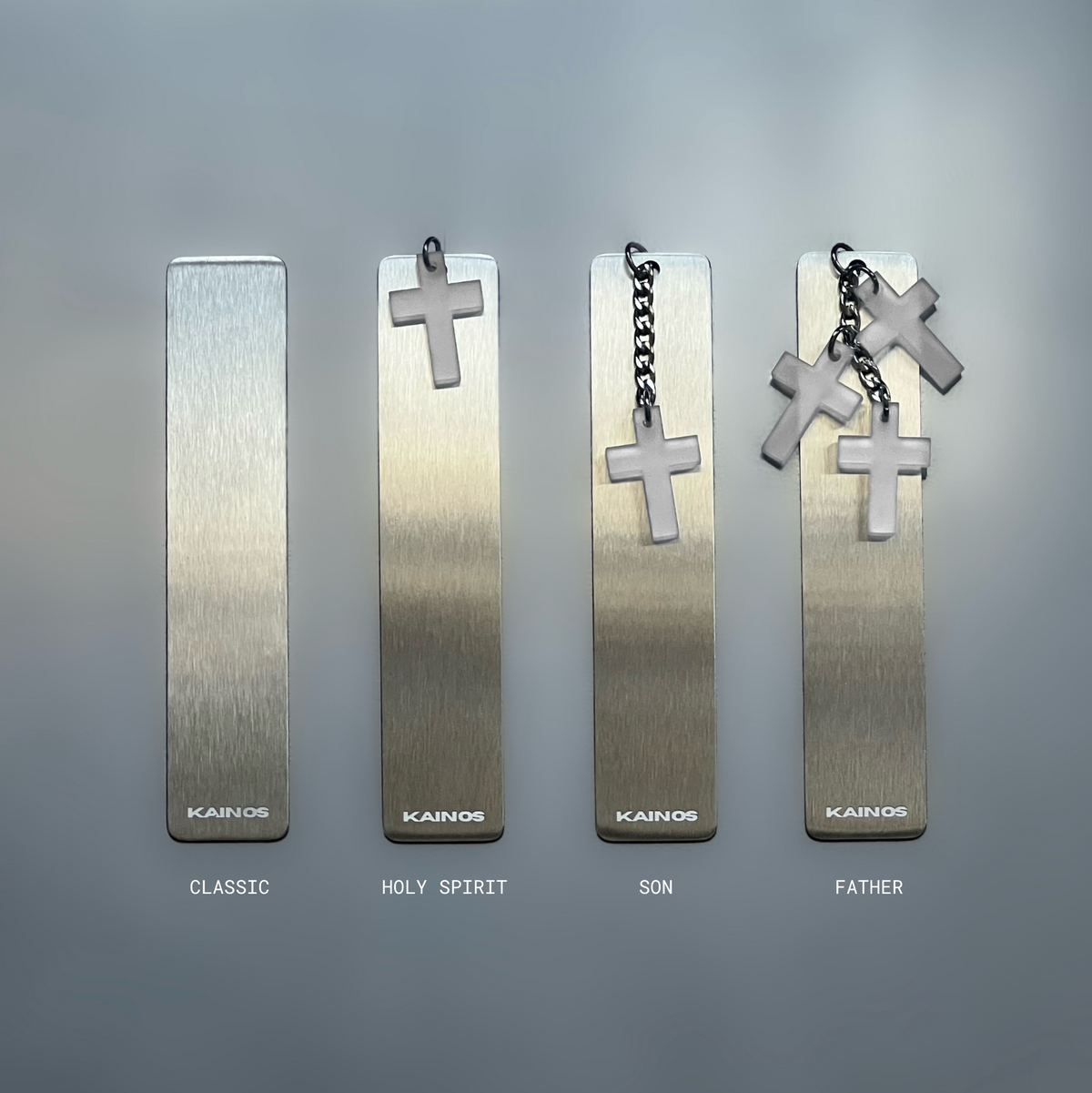 Four rectangular metallic bookmarks or tags with hanging cross charms are arranged on a smooth gray surface. From left to right, the designs are labeled Classic, Holy Spirit, Son, and Father, with the word 'KAINOS' visible near the bottom of each piece.