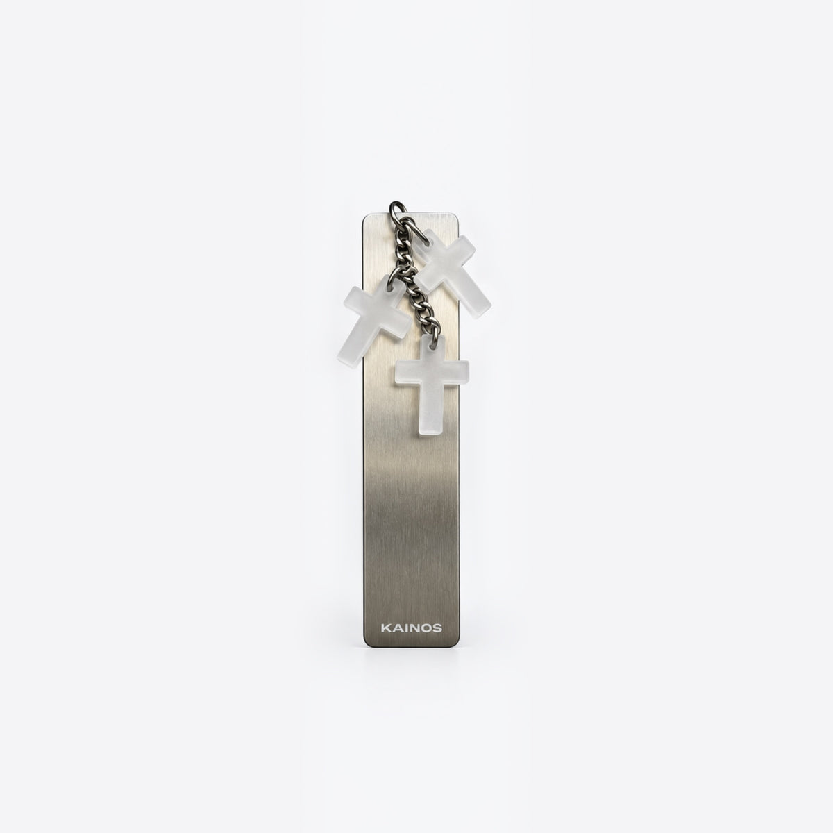 A slim rectangular metallic bookmark or card holder with a brushed silver finish, featuring three hanging translucent white crosses on a short chain and the brand name 'KAINOS' near the bottom, shown on a clean white background.