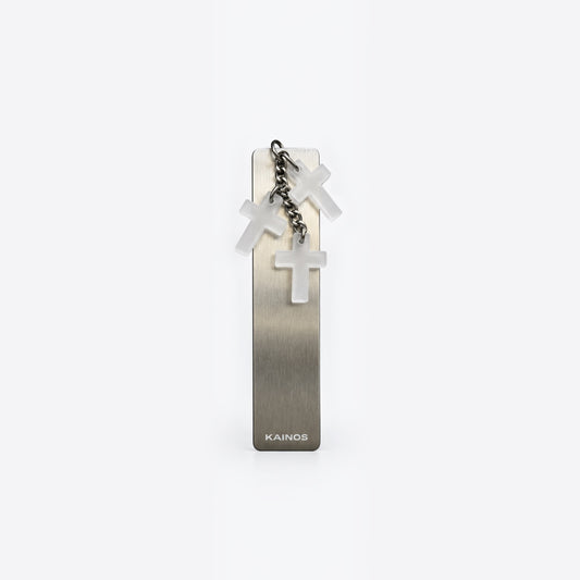 A slim rectangular metallic bookmark or card holder with a brushed silver finish, featuring three hanging translucent white crosses on a short chain and the brand name 'KAINOS' near the bottom, shown on a clean white background.