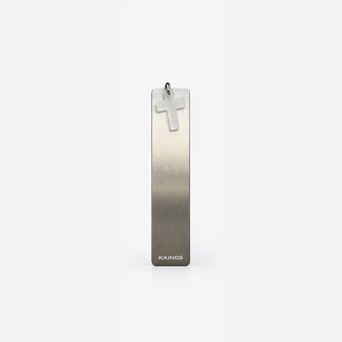 A vertical rectangular brushed metal pendant with a small cross charm at the top and the brand name KAINOS near the bottom, shown on a clean light gray background.