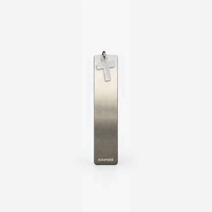 A vertical rectangular brushed metal pendant with a small cross charm at the top and the brand name KAINOS near the bottom, shown on a clean light gray background.
