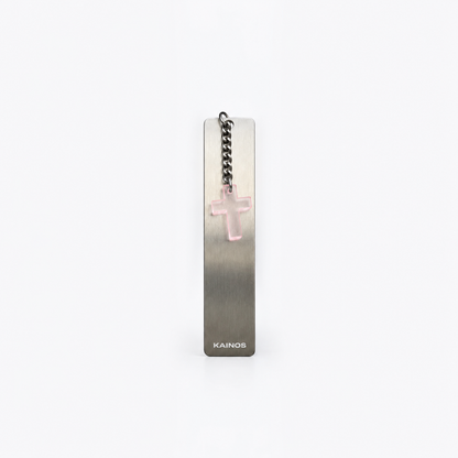 A slim rectangular metal bookmark with a brushed silver finish, featuring a small pink translucent cross charm on a black chain and the brand name 'KAINOS' at the bottom, set against a plain white background.