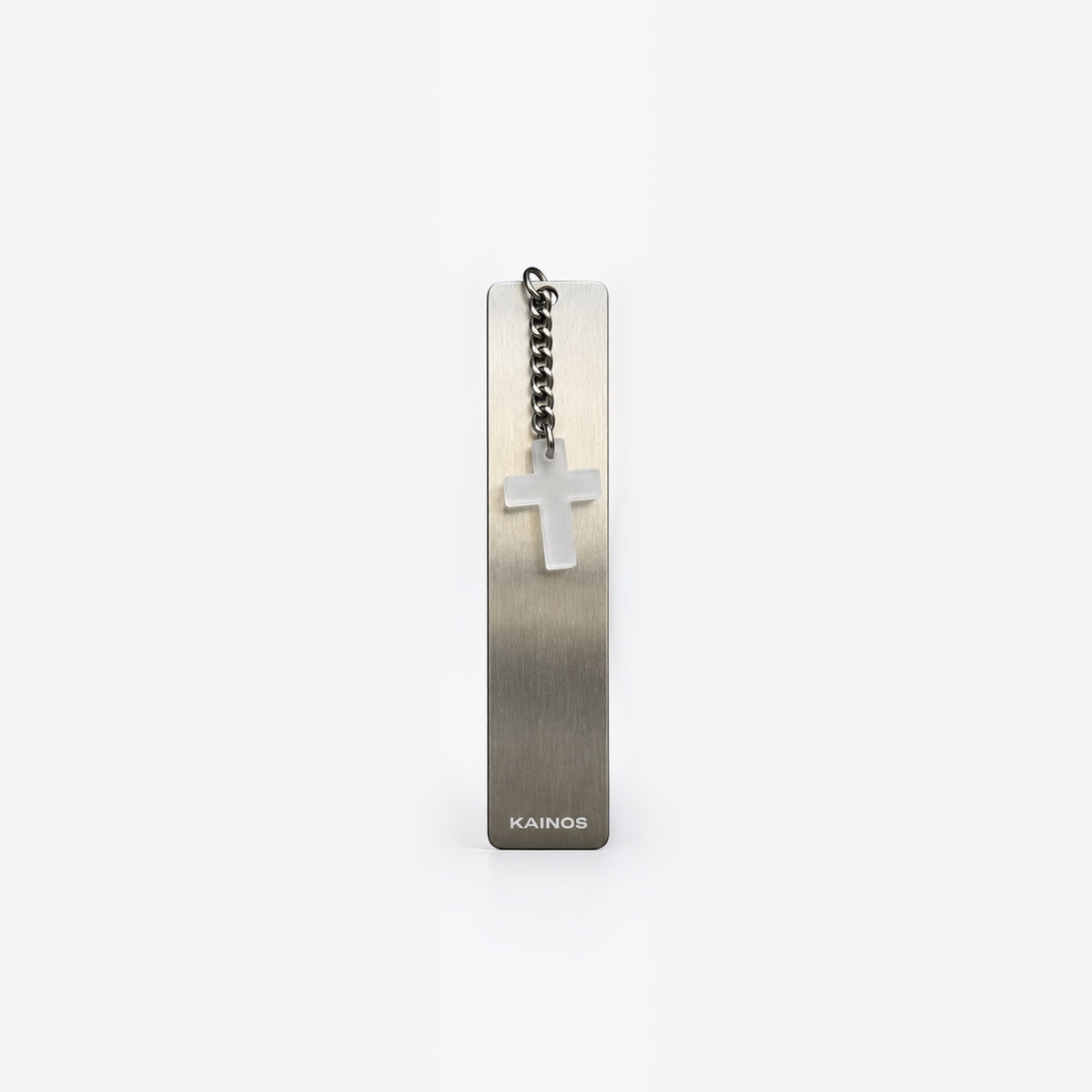 A slim rectangular metal bookmark or tag with a brushed silver finish, featuring a small white cross charm on a short chain and the brand name KAINOS near the bottom, shown on a light gray background.