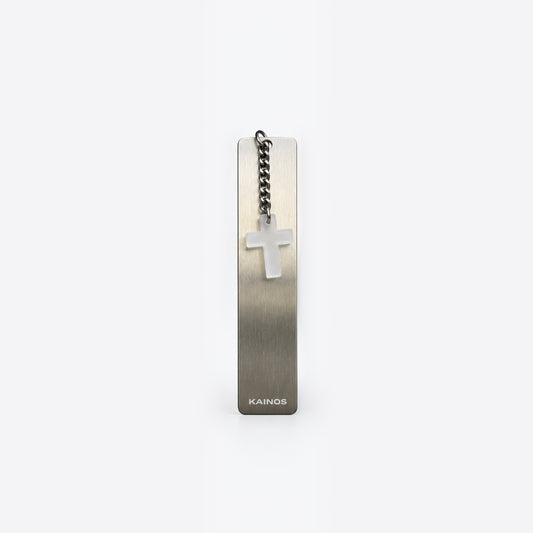 A slim rectangular metal bookmark or tag with a brushed silver finish, featuring a small white cross charm on a short chain and the brand name KAINOS near the bottom, shown on a light gray background.