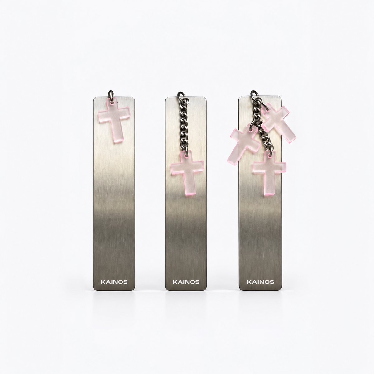 Three Kainos rectangular metal bookmarks or tags with brushed silver-gray finish, each hanging from a chain with a translucent pink cross charm, shown on a clean white background.