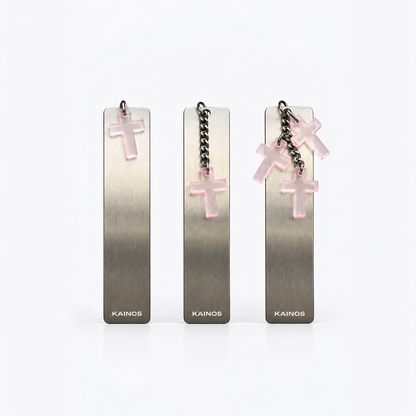 Three Kainos rectangular metal bookmarks or tags with brushed silver-gray finish, each hanging from a chain with a translucent pink cross charm, shown on a clean white background.