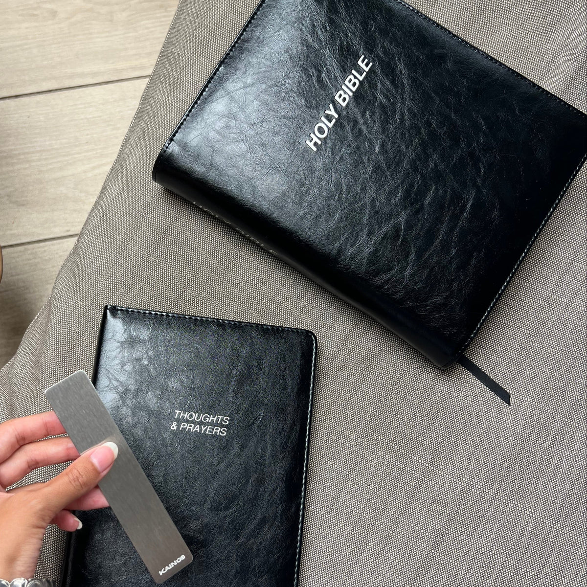 Two black textured leather-bound books, one labeled HOLY BIBLE and the other THOUGHTS & PRAYERS, laid on a gray fabric surface, with a hand holding a metal ruler next to the Thoughts & Prayers book.