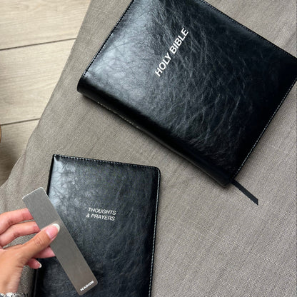 Two black textured leather-bound books, one labeled HOLY BIBLE and the other THOUGHTS & PRAYERS, laid on a gray fabric surface, with a hand holding a metal ruler next to the Thoughts & Prayers book.