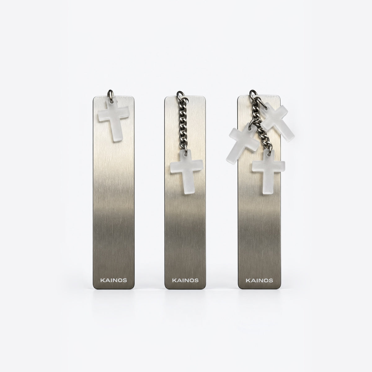 Three Kainos rectangular metal pendant earrings with brushed silver gradients, each featuring a small white cross charm and hanging chain, displayed on a clean white background.
